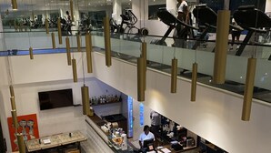 Un restaurante moderno con mesas y sillas de madera, una barra con un empleado, y una escalera visible que lleva a un nivel superior.