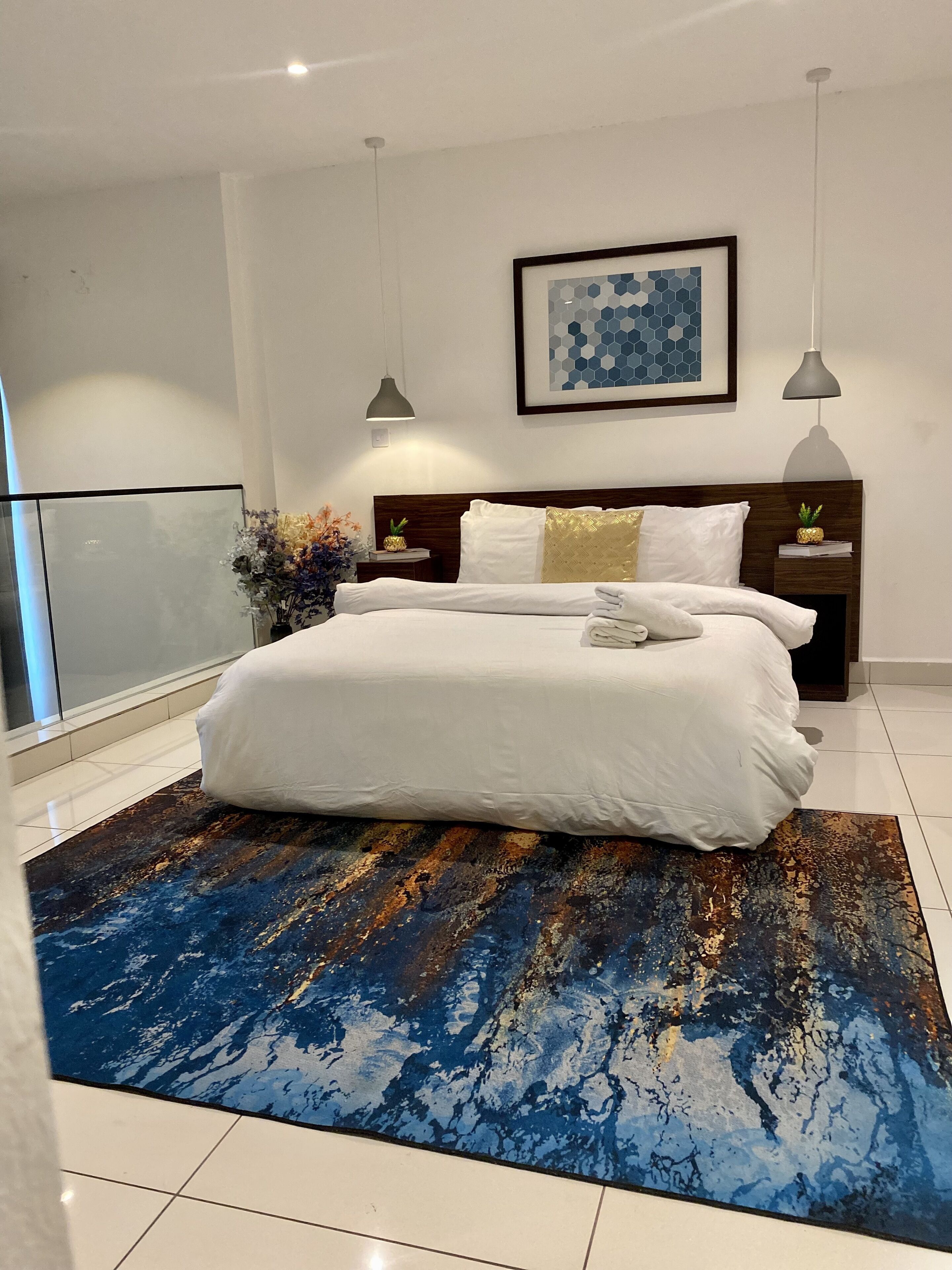 A modern bedroom with a large bed, a blue abstract rug, a framed artwork on the wall, and a nightstand with a pineapple decoration.
