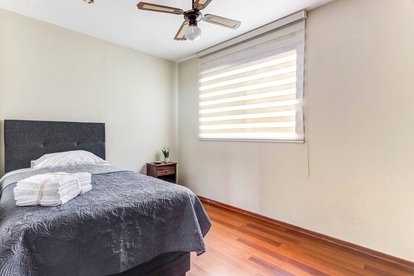 A bedroom with a bed, a nightstand, a ceiling fan, and a window with blinds.