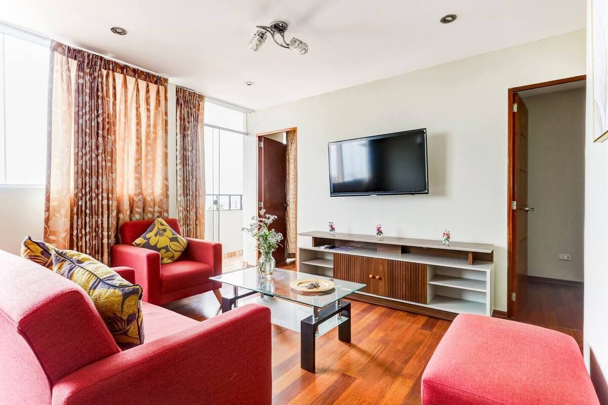 A modern living room with a flat-screen TV, red sofas, a glass coffee table, and a wooden entertainment unit.