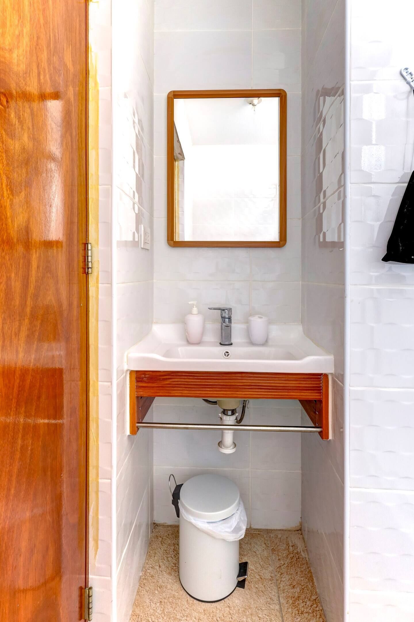 A bathroom with a wooden door, white tiled walls, a white sink with a mirror above it, a toilet, and a trash bin.