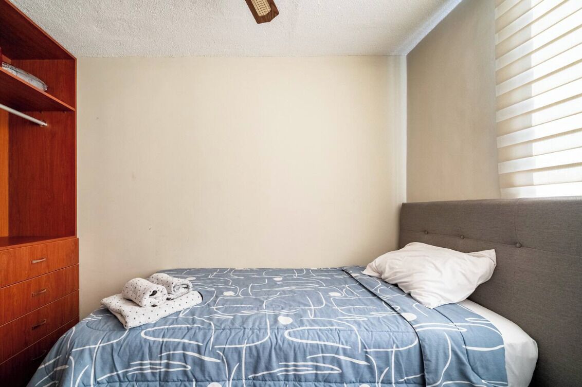 Een slaapkamer met een bed, een grijs gestoffeerd hoofdeinde, een houten garderobe en een raam met zonwering.