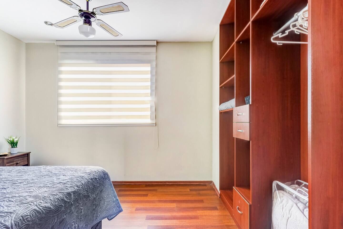 A bedroom with a bed, wooden wardrobe, ceiling fan, and a window with blinds.