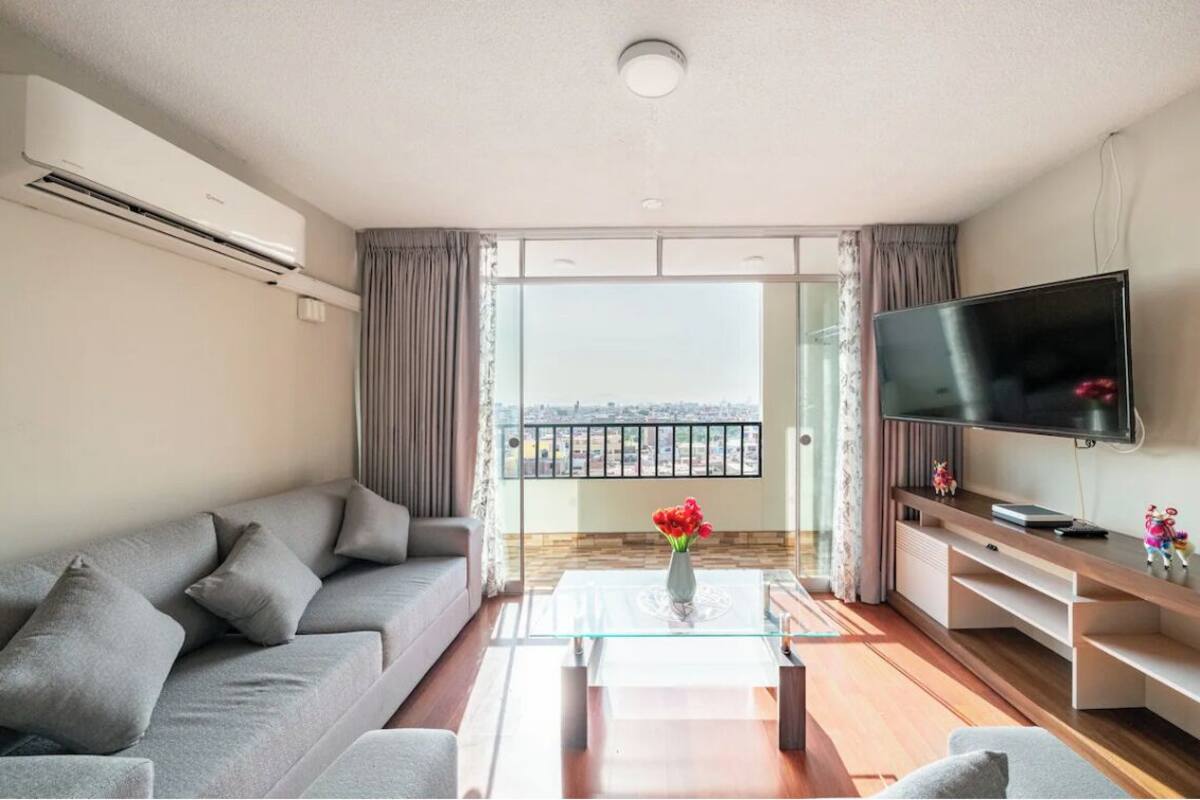A modern living room with a grey sofa, a glass coffee table, a flat-screen TV, and a balcony with a view.
