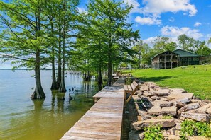 A wooden dock extending into a lake with trees on one side and a lodge with a porch on the other. - Lakefront House w/ Deck & Fire Pit (Vicksburg)