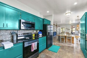 Una cocina con gabinetes color turquesa, un refrigerador negro y un microondas. Hay un comedor con una mesa y sillas de madera.