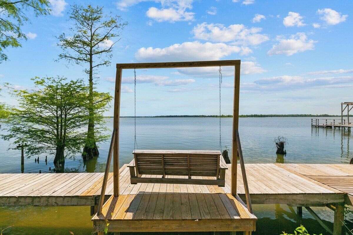 Une balançoire en bois surplombe un lac paisible, avec un quai en bois et des arbres en arrière-plan.