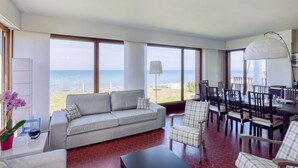 Amplia sala de estar con un gran ventanal con vistas al mar, un sofá, una mesa de comedor con sillas y una lámpara.