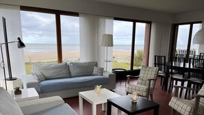 Una sala de estar moderna con un ventanal que da a una playa, un sofá, una mesa de centro y una zona de comedor con sillas.