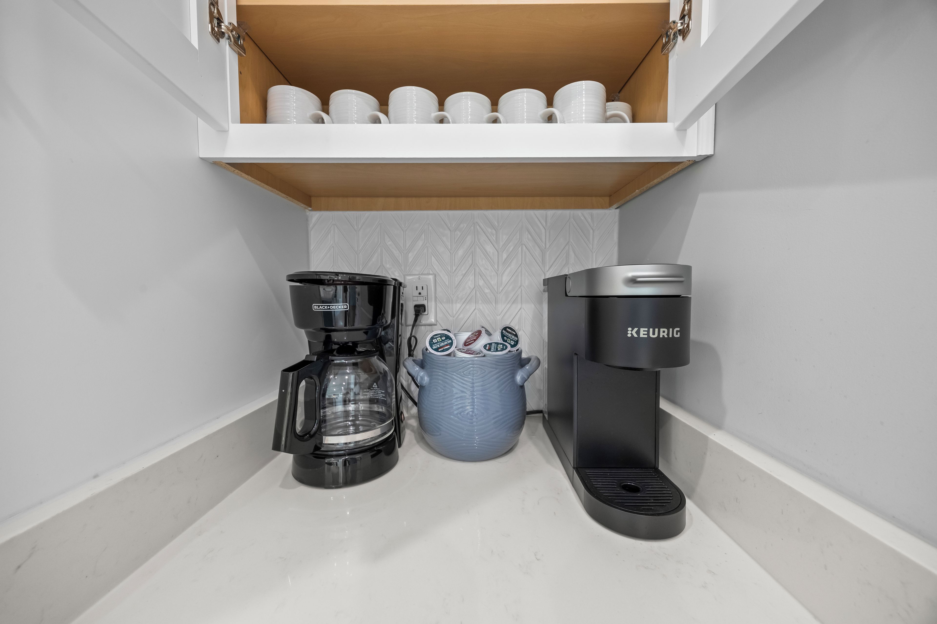 Una cocina con cafetera, jarra de café y estante con tazas blancas.