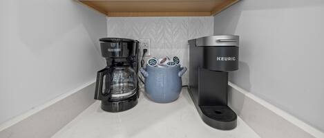 Una cocina con cafetera, jarra de café y estante con tazas blancas.