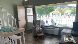 A room with a dining table, a chair, a sofa, a window with a view of the street, and a sign on the door.