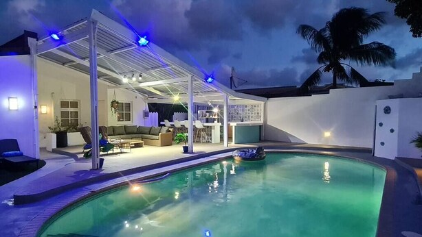 Une piscine avec un patio couvert, un salon doté de sièges et un palmier en arrière-plan.