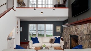 A spacious living area with a high ceiling, a stone fireplace, and a large window overlooking a scenic view.