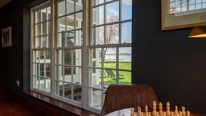 A room with a large window overlooking a grassy area and a body of water. A chessboard is placed on a table in the foreground.