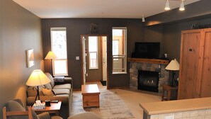 Ein Wohnzimmer mit Sofa, Hocker, Couchtisch und Kamin. Über dem Kamin hängt ein Fernseher und auf einem Beistelltisch steht eine Lampe.