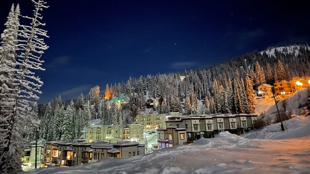 Eine schneebedeckte Bergstadt bei Nacht mit beleuchteten Gebäuden und freiem Blick auf den umliegenden Wald.
