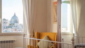 A room with a bed, a yellow armchair, and a view of a cityscape including a large dome building. - Saint Peter View - Saint Peter View 2 pax (Rome)