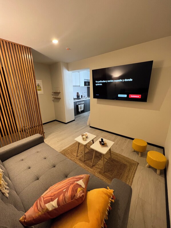 A modern living room with a grey sofa, a flat-screen TV mounted on the wall, and a kitchen in the background. - Ocean view! Great location, ideal for couples or small families (Barranco)