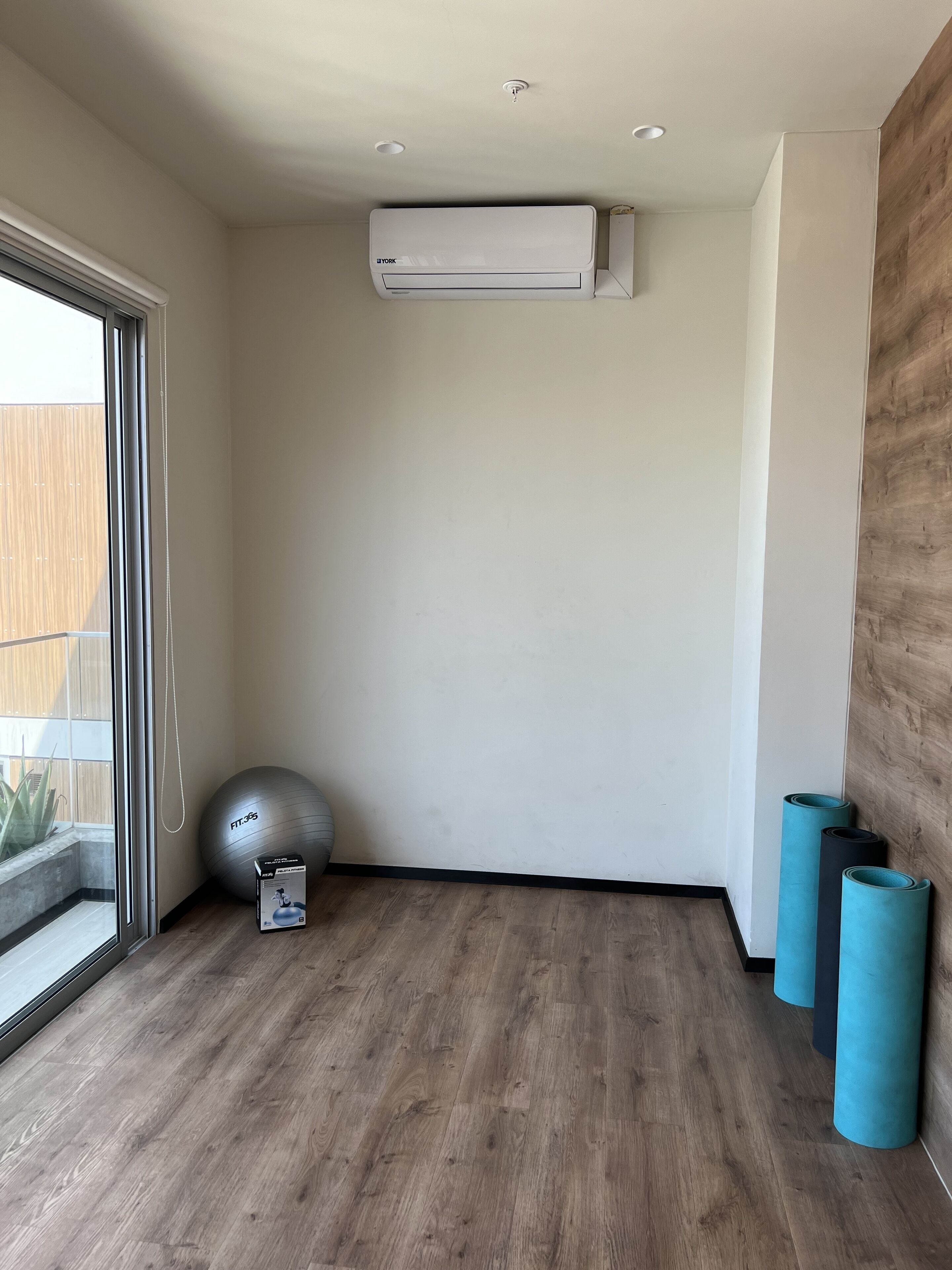 Une pièce dotée d’une grande fenêtre, d’un climatiseur, d’un ballon d’exercice gris et de trois tapis de yoga enroulés.