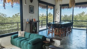 Una sala de estar moderna con un sofá verde, un comedor con una mesa redonda y sillas, y ventanas amplias que ofrecen vistas a zonas verdes y un edificio.