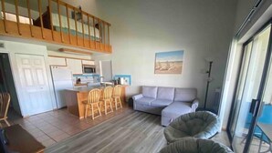 Uma sala de estar ampla com sofá, dois pufes, mesa de jantar com cadeiras, cozinha e varanda com guarda-corrimão.