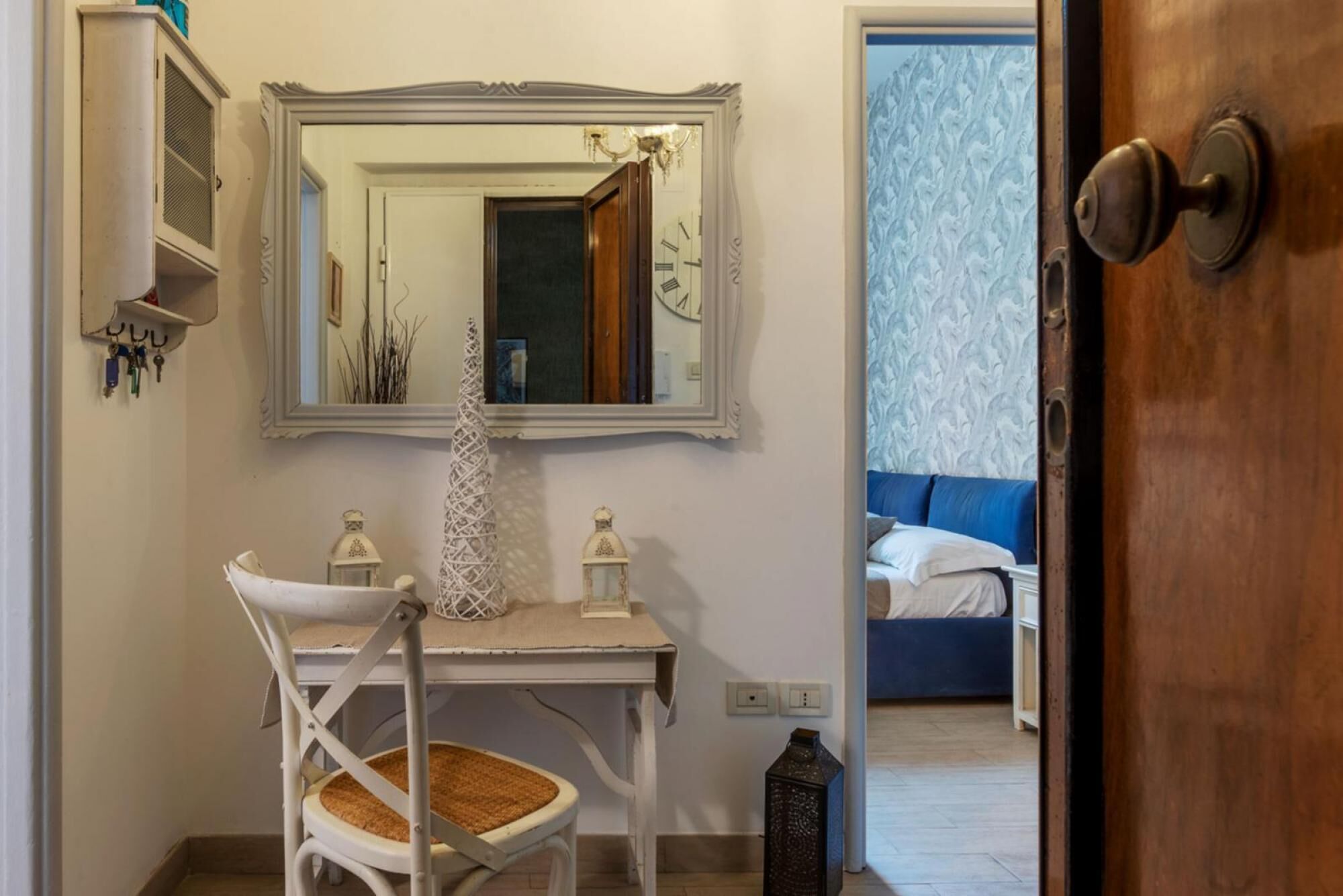 A bathroom with a white vanity, a mirror, and a chair. There is a door leading to a bedroom with a blue bed and a patterned wall.
