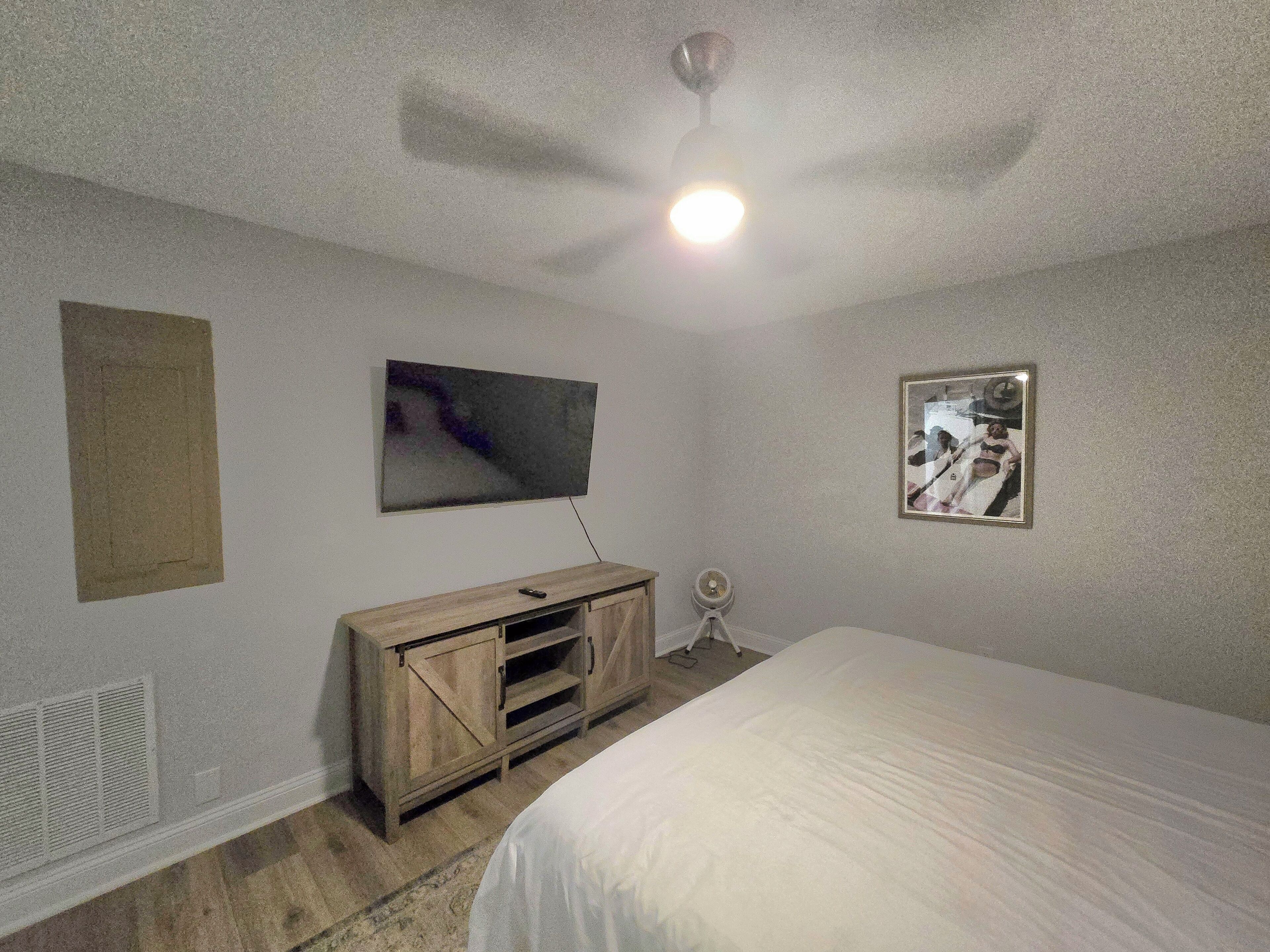 A bedroom with a bed, a TV mounted on the wall, a wooden cabinet, and a small table with a lamp.