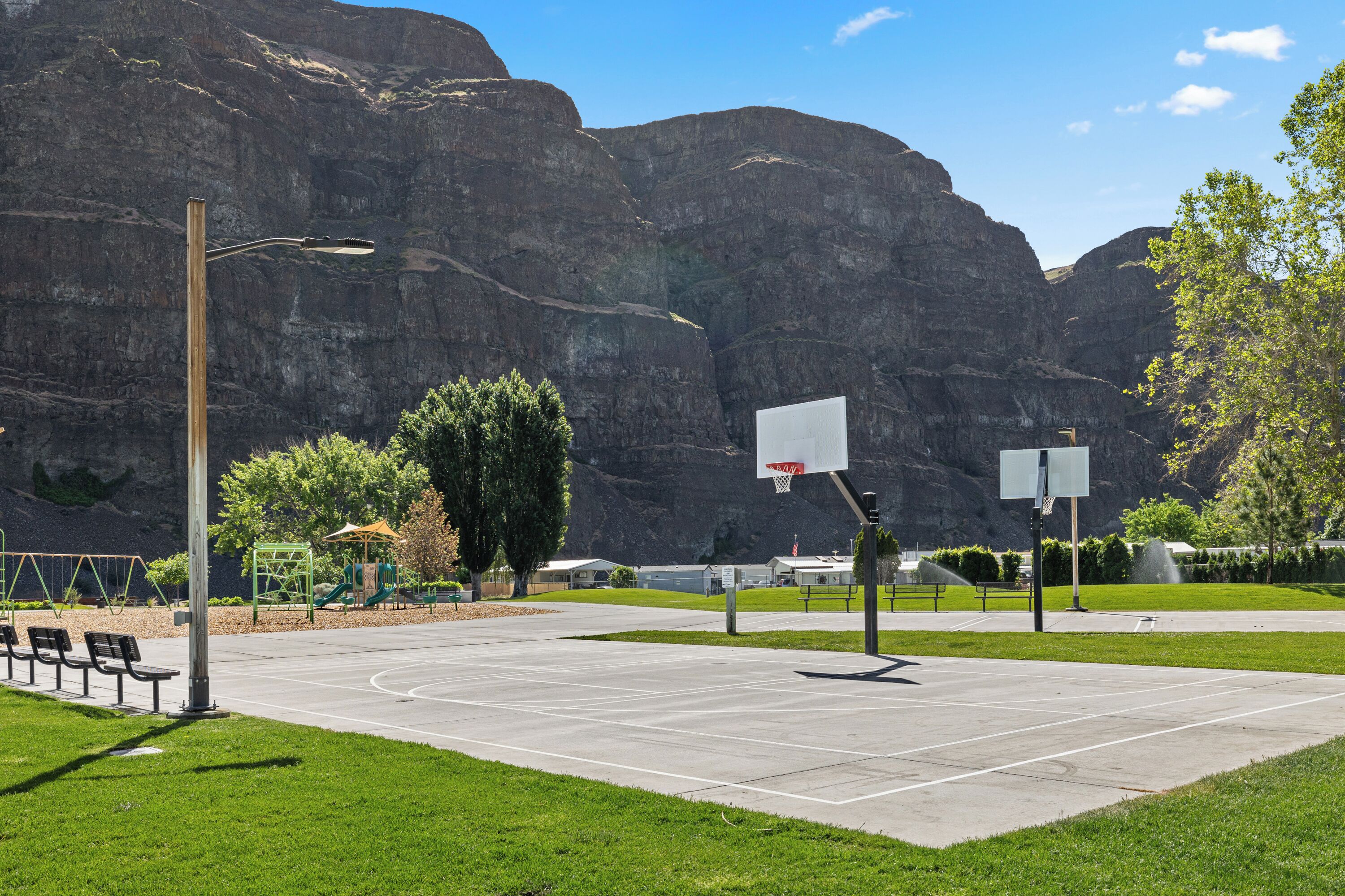 An outdoor basketball court with a green playground and benches, set against a mountainous backdrop.