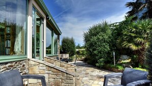 A stone patio with wicker furniture, a stone wall, and a glass door leading to a room. - Simple Bonheur (Penvénan)