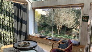 A sunlit room with a large window, a round table with a glass top, a cushioned bench, and a chair with a patterned cushion. - Simple Bonheur (Penvénan)