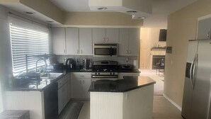 Een moderne keuken met witte kasten, een centraal kookeiland, roestvrijstalen apparatuur en een groot raam met zonwering.