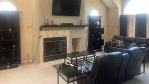 Een ruime woonkamer met een open haard, een flatscreen televisie aan de muur en een plafondventilator.