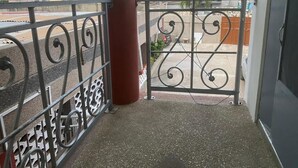 Un balcón con una reja metálica, un pilar rojo y vistas a una calle con edificios y palmeras.