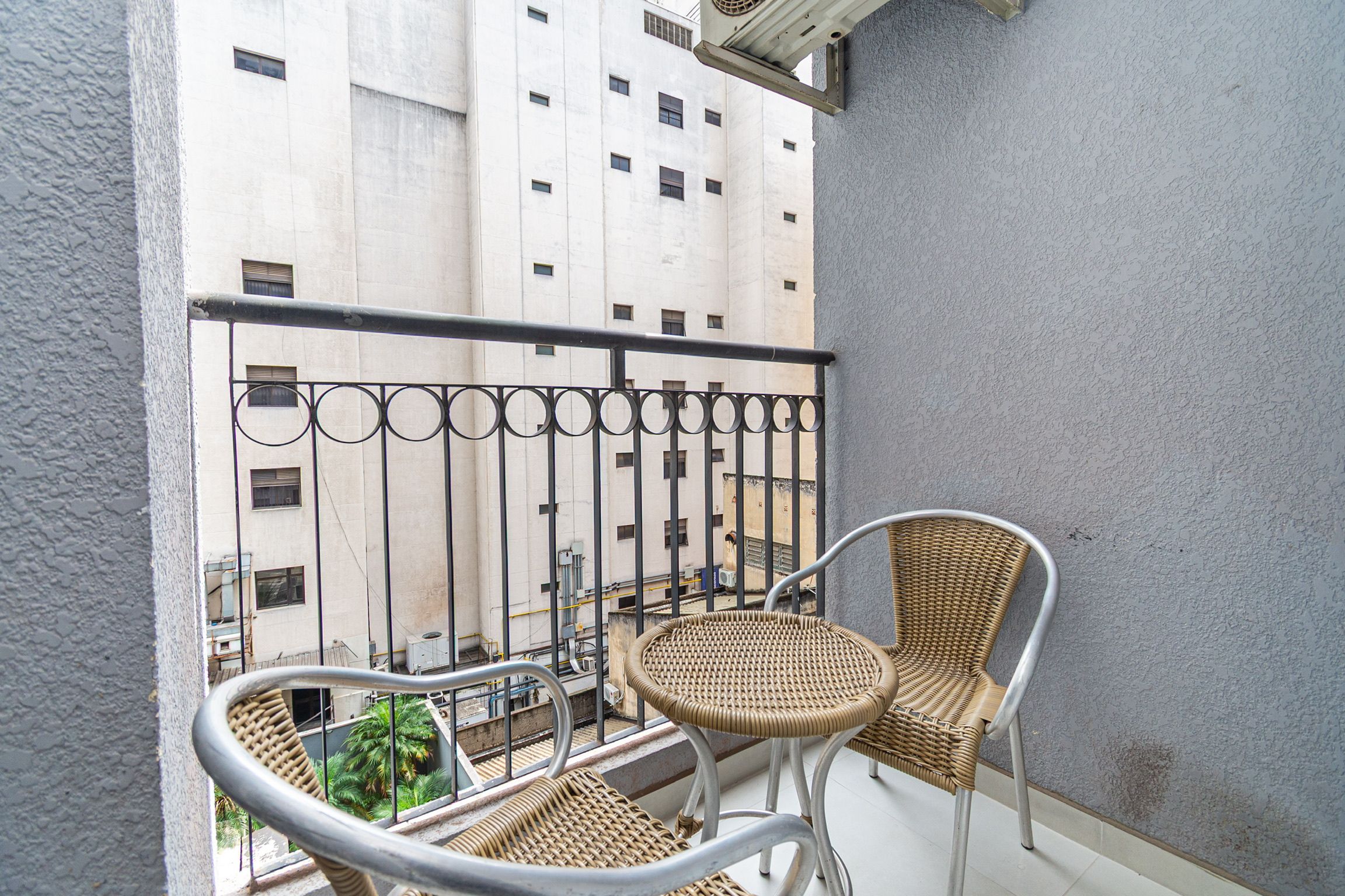 A balcony with two wicker chairs and a small table, overlooking a building with multiple windows.