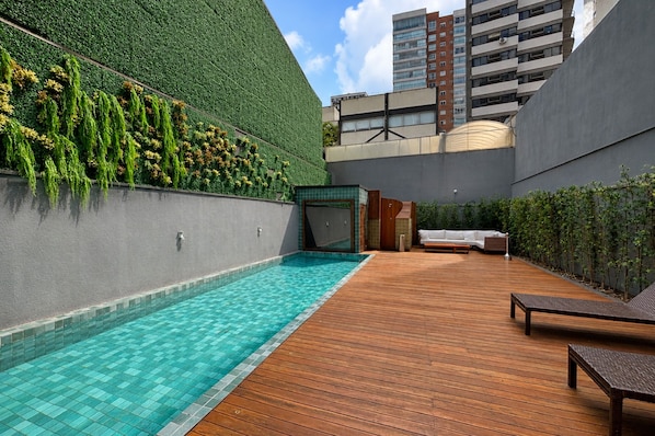 Un área de piscina en la azotea con cubierta de madera, un muro verde con plantas y vistas de edificios circundantes.