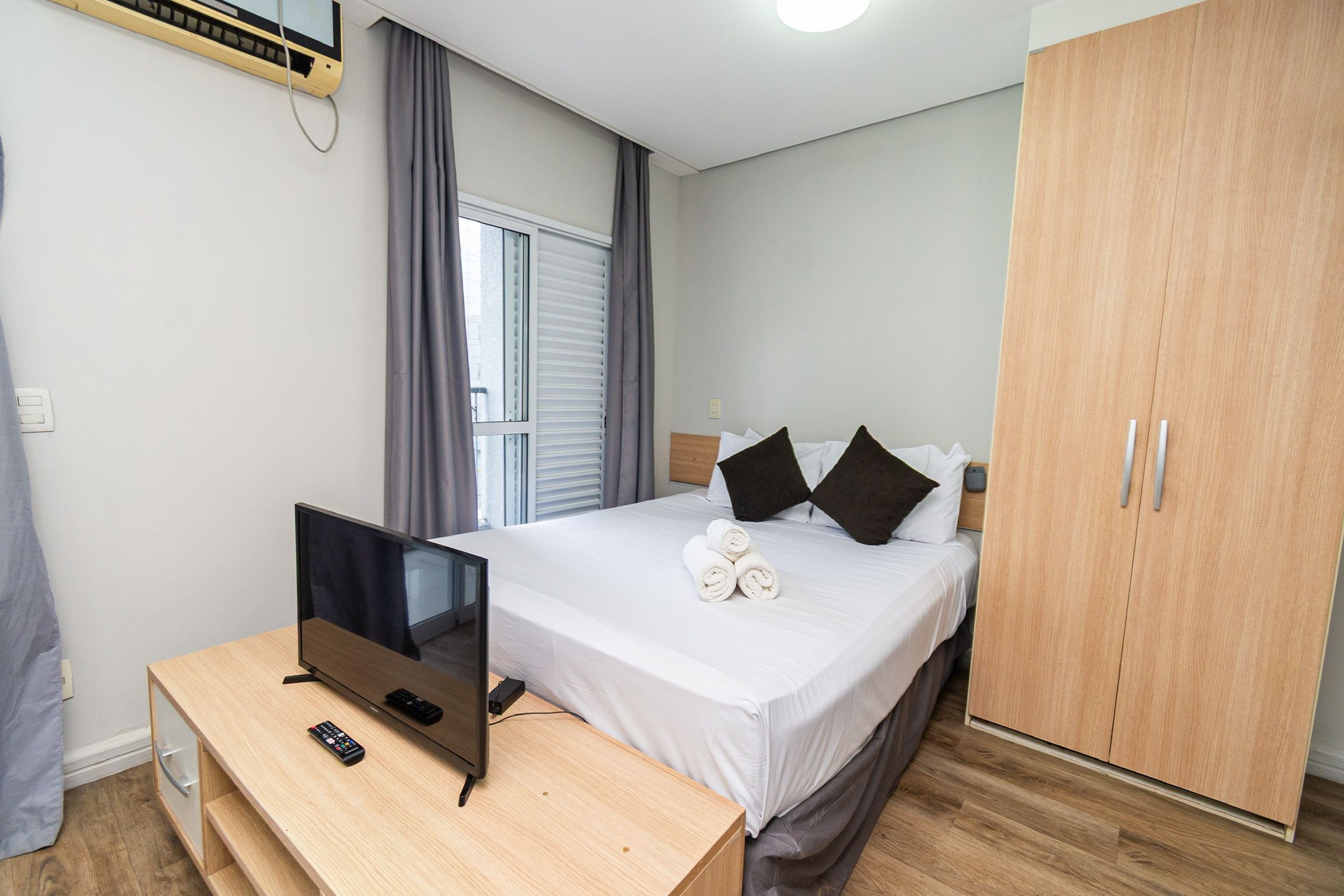 Ein Hotelzimmer mit Bett, Schreibtisch mit Fernseher und Fernbedienung, Kleiderschrank und einem Vorhangfenster.