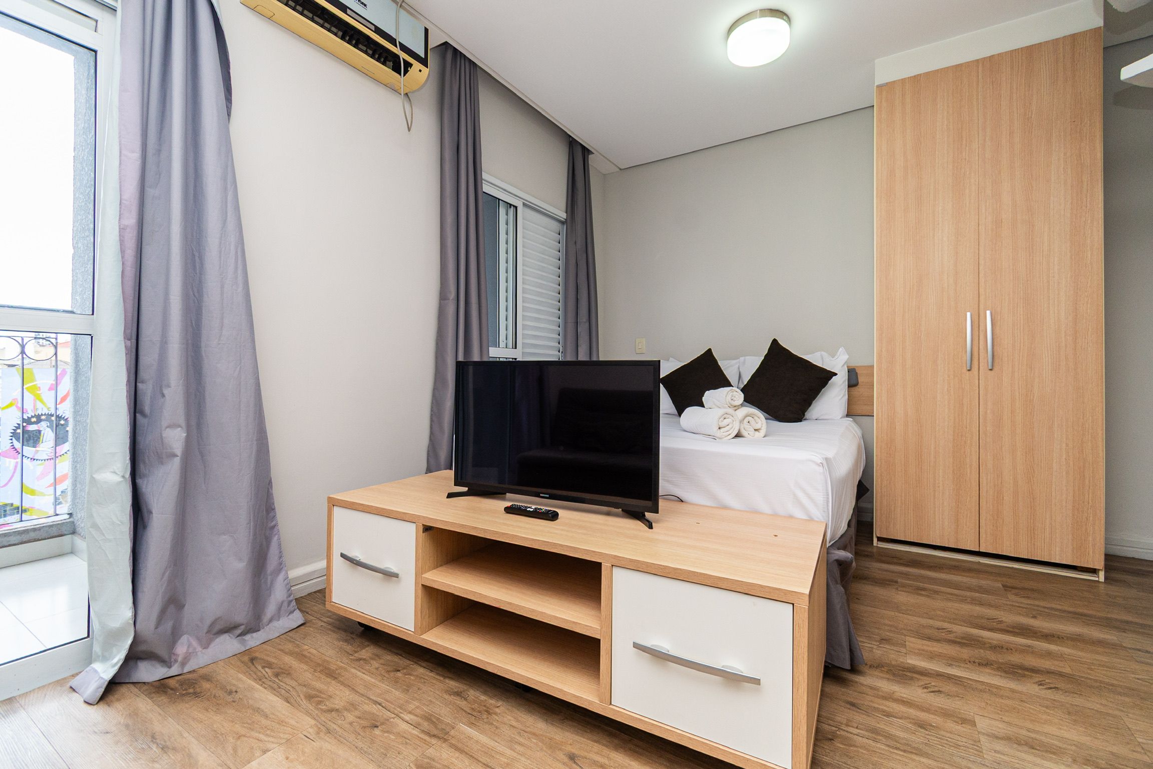 Un dormitorio con cama, televisor sobre un soporte de madera, armario y ventana con cortinas.