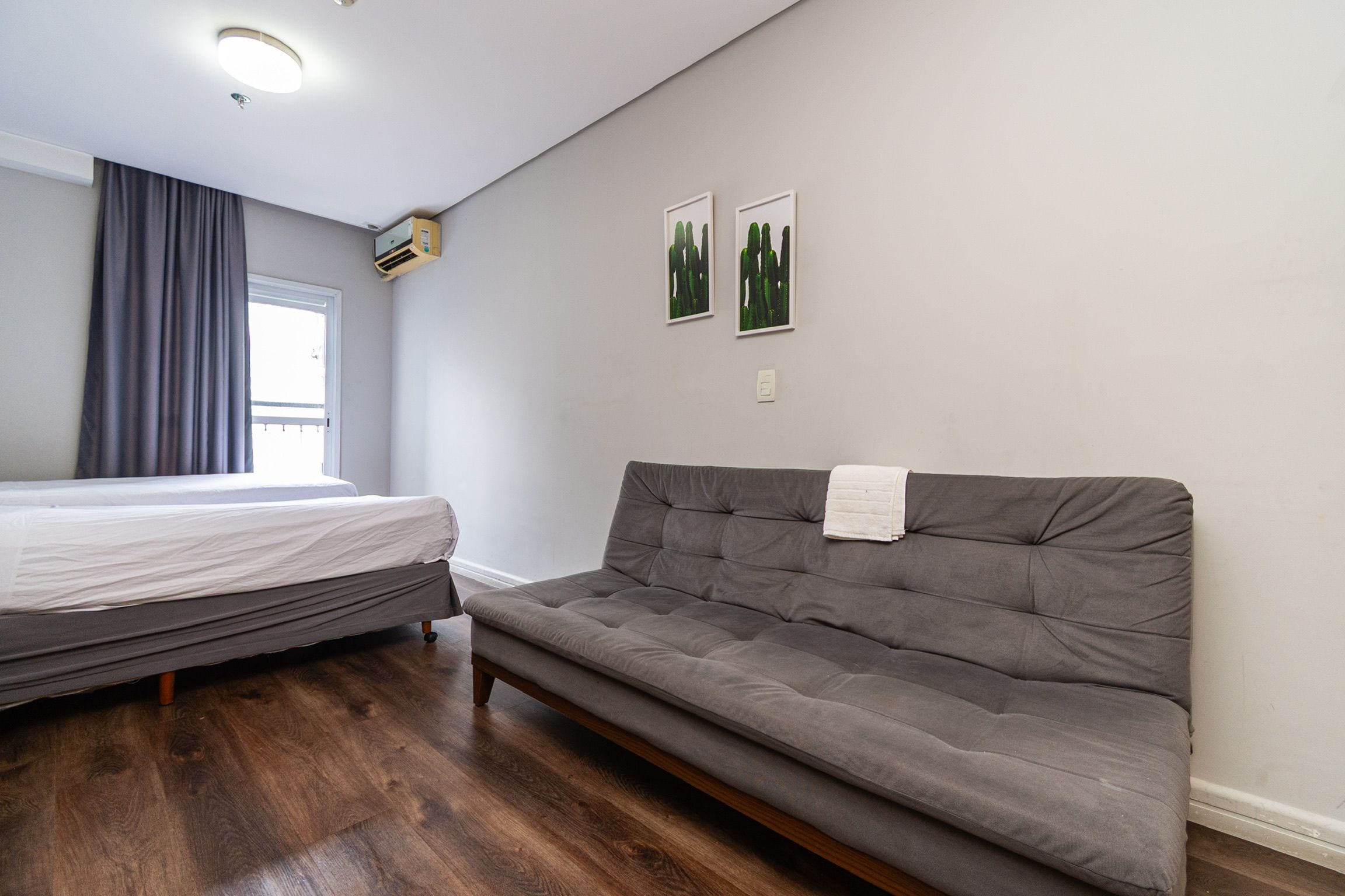 A room with a bed, a sofa, a window with curtains, and two wall-mounted pictures.