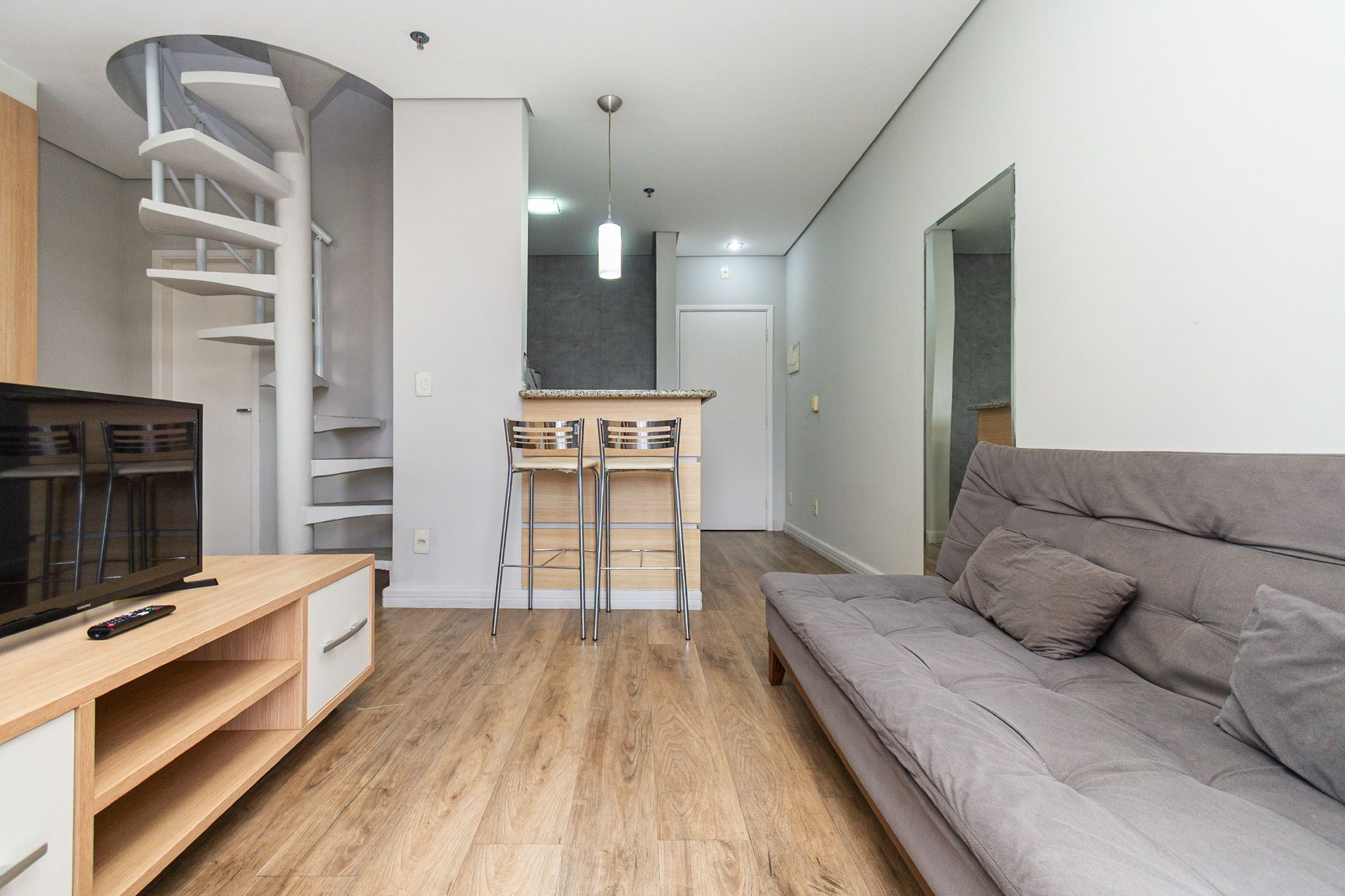 Una sala moderna con un sofá gris, un televisor, un escritorio de madera y una zona de cocina con mostrador y taburetes.