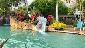Una piscina con tobogán de agua, rodeada de plantas tropicales y una cascada.