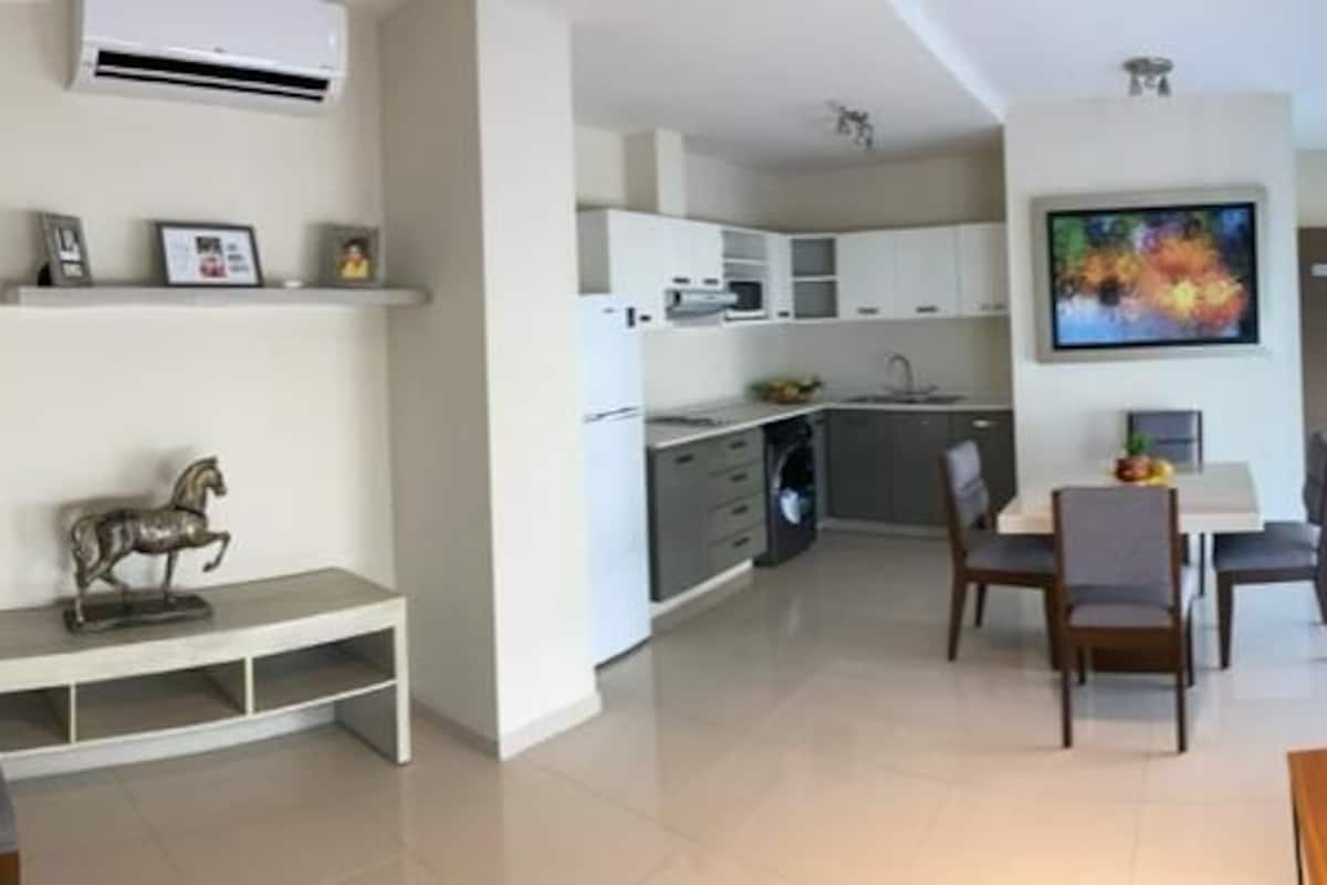 A modern living room with a grey sofa, a dining table with chairs, and a kitchen area in the background.