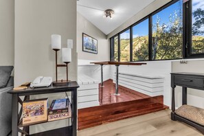 A modern interior with a staircase leading to a balcony, a desk with a phone and board game, and a view of trees through large windows.