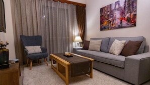 Een woonkamer met een grijze bank, een blauwe fauteuil, een houten salontafel en een schilderij van een stadsgezicht.