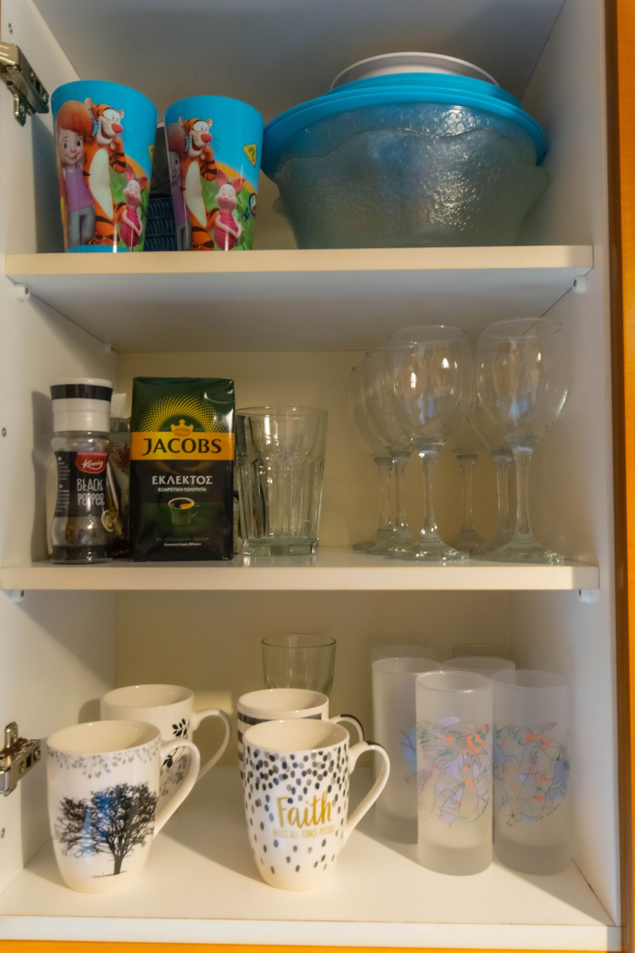A shelf with various items including a pack of Jacobs black pepper, clear wine glasses, and decorative mugs.