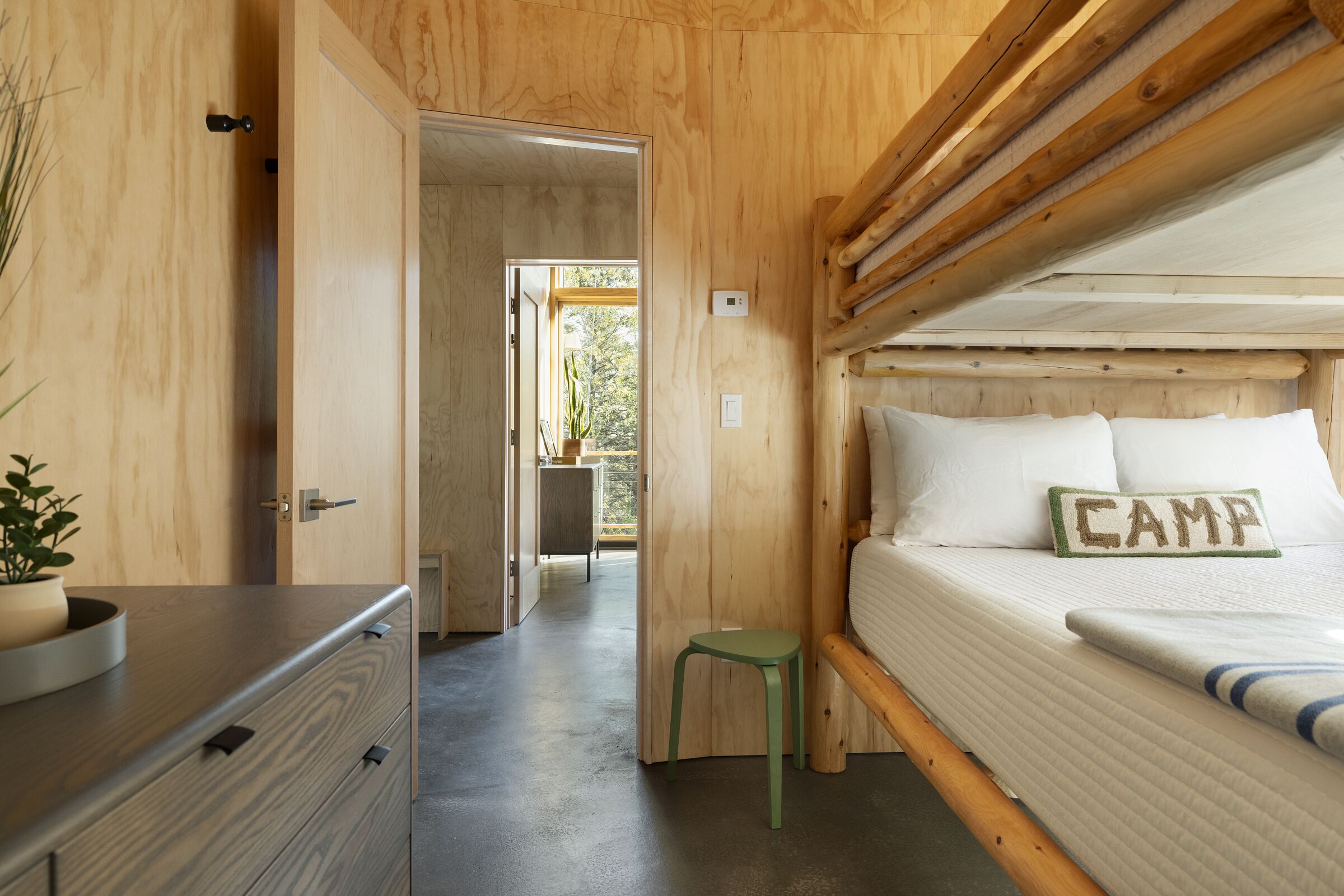 A wooden cabin interior with a bed labeled "CAMP," a small stool, and a dresser with a plant.