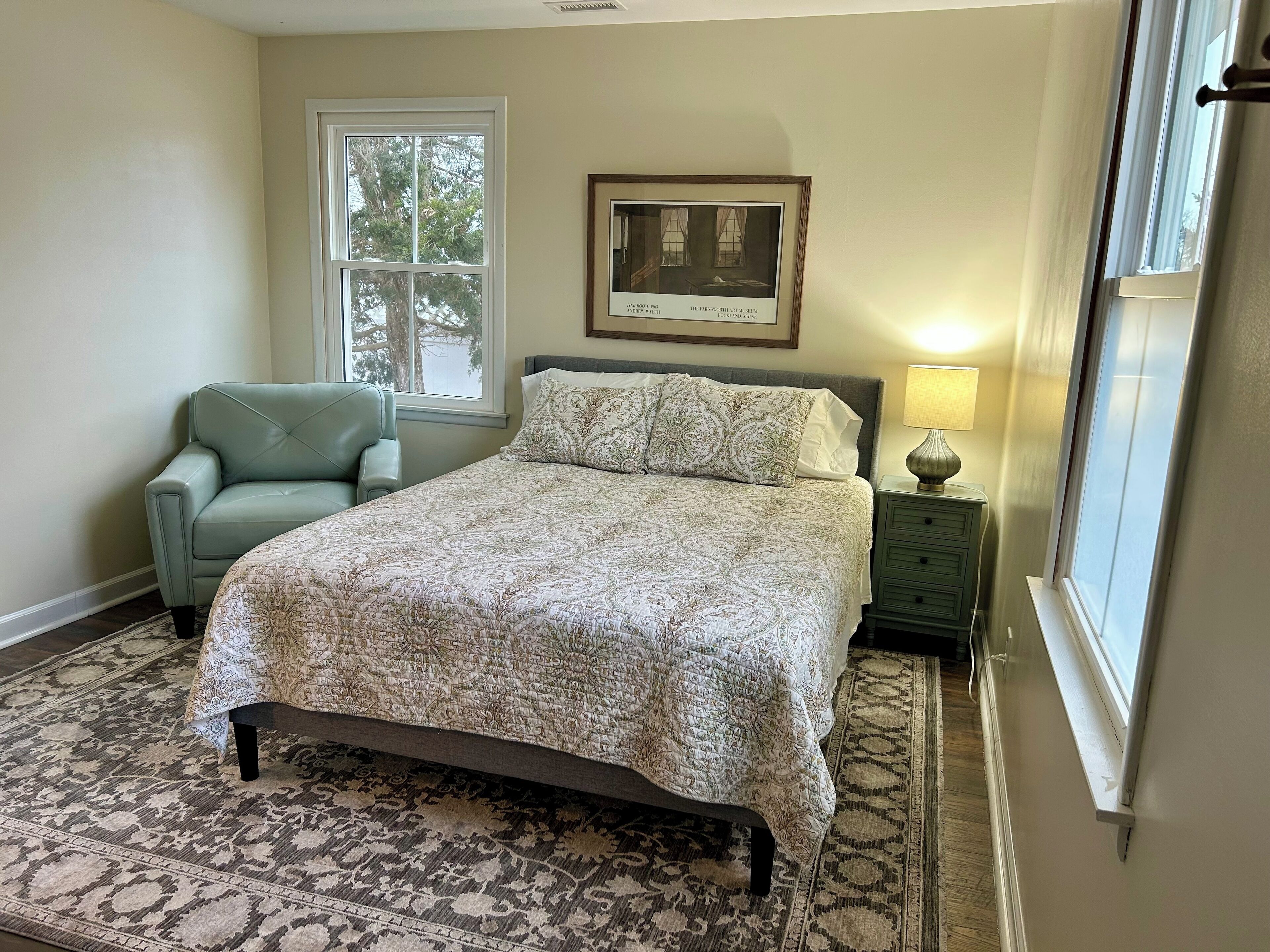 A bedroom with a large bed, a chair, a nightstand with a lamp, and a framed picture on the wall.