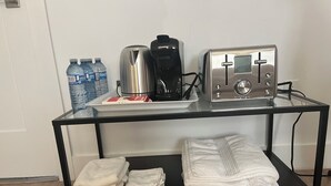 Une table avec un plateau en verre, une machine à café, un grille-pain et des bouteilles d’eau. Des serviettes pliées sont posées sur l’étagère inférieure.