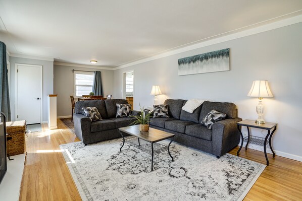 A living room with two grey sofas, a wooden coffee table, and a patterned rug. There are two lamps on side tables, a painting on the wall, and a door leading to another room.