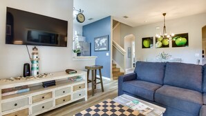 Una sala de estar con un televisor de pantalla plana grande, un sofá azul, una mesa de centro con mantel individual a cuadros y una estantería con libros y objetos decorativos.